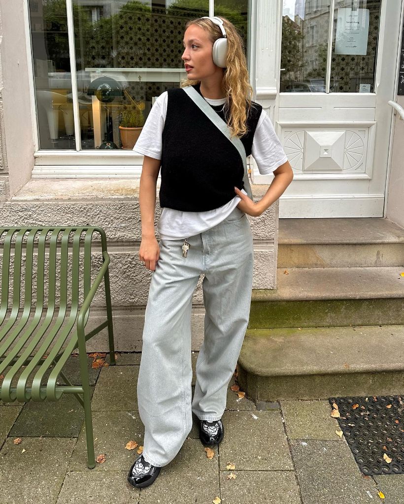 White tee layered under black knit vest with light wash wide-leg jeans, headphones, and patterned loafers