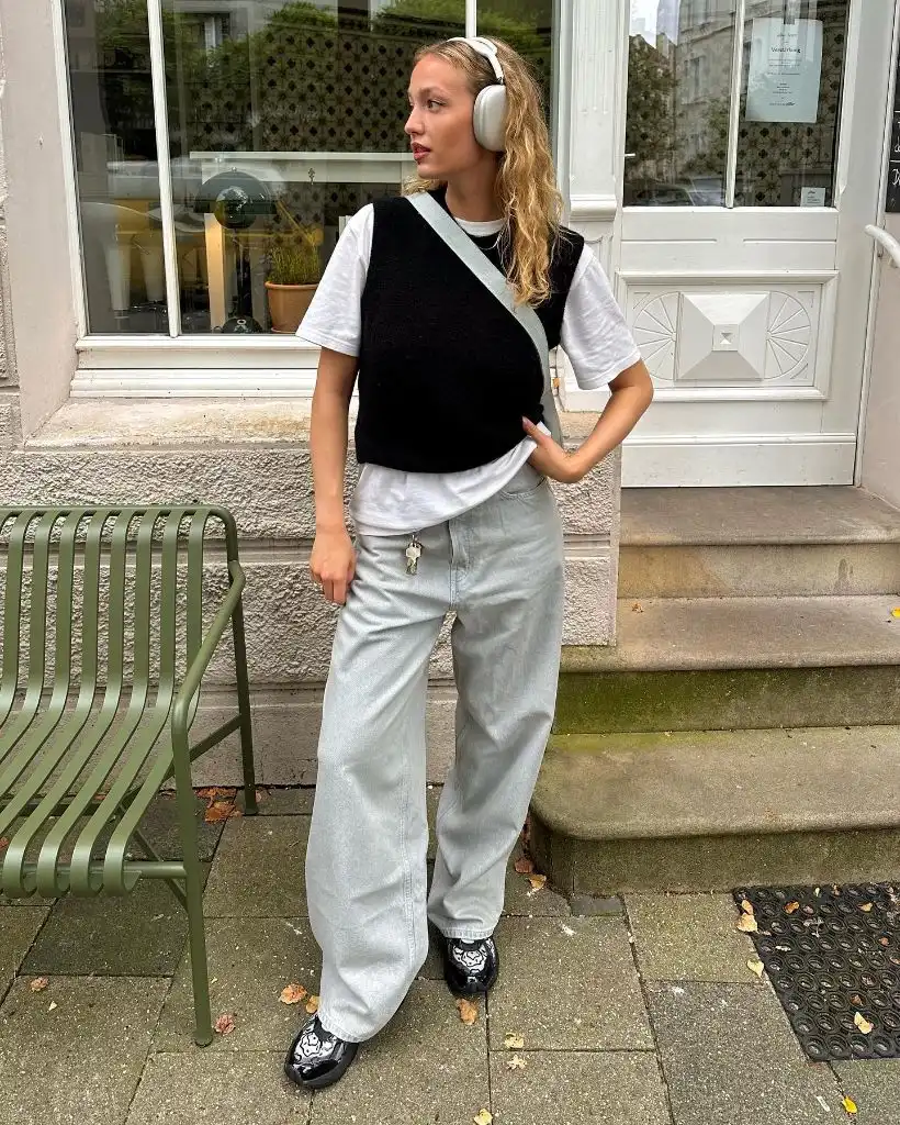 White tee layered under black knit vest with light wash wide-leg jeans, headphones, and patterned loafers