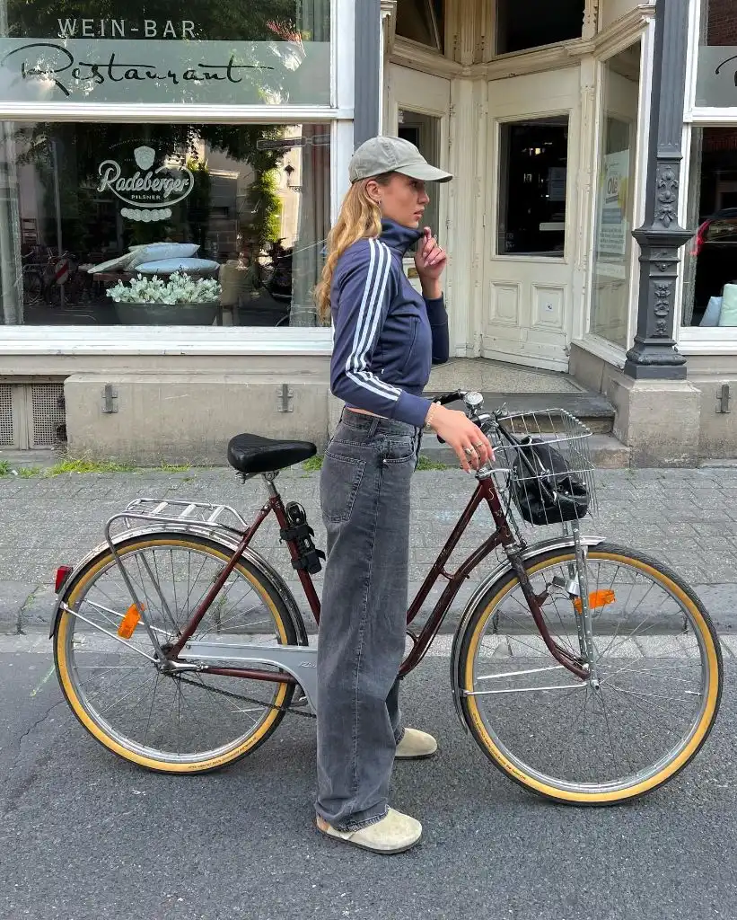 Navy Adidas track jacket with grey wide-leg jeans, grey cap, and cream slip-on shoes next to bicycle
