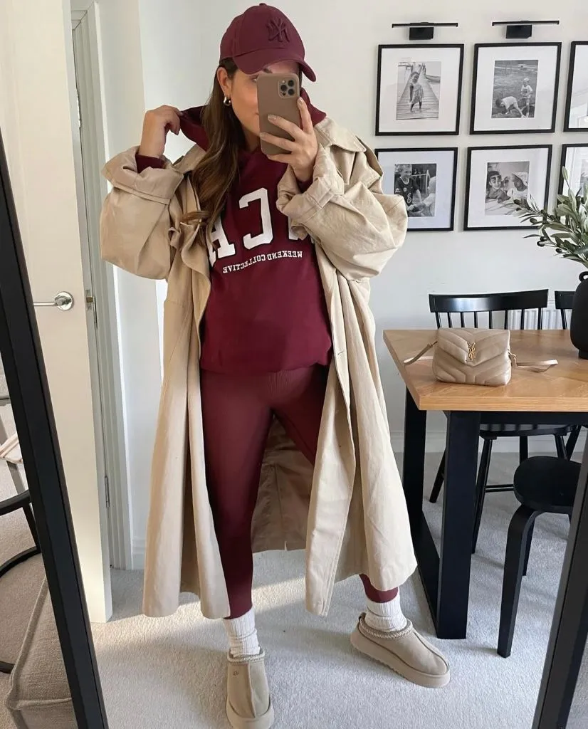 Mirror selfie of pregnant woman in burgundy hoodie and leggings, cream trench coat, UGG slides, and YSL bag