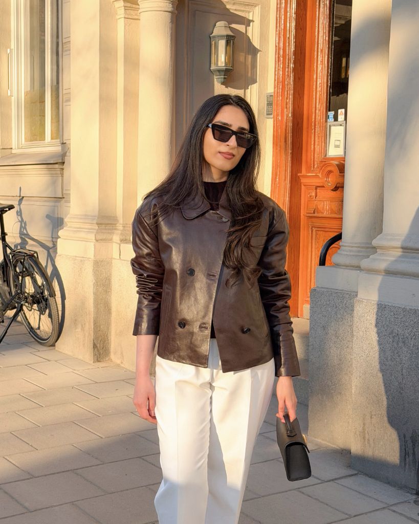 Dark brown leather jacket with white trousers and small bag on European street