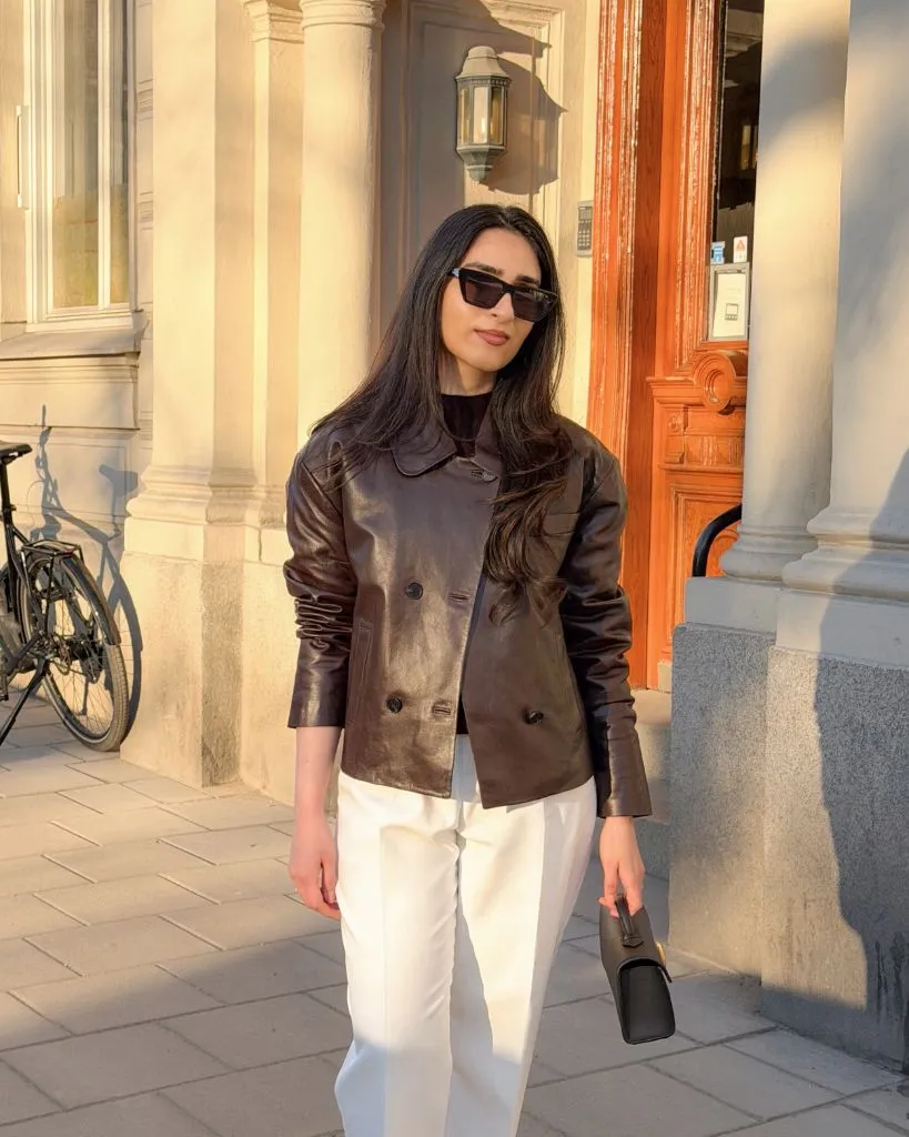 Dark brown leather jacket with white trousers and small bag on European street