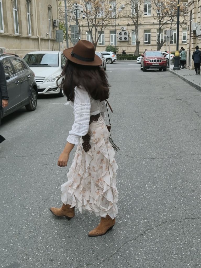 Walking shot of ruffled floral skirt with white blouse, cowboy boots, brown hat, and crossbody bag