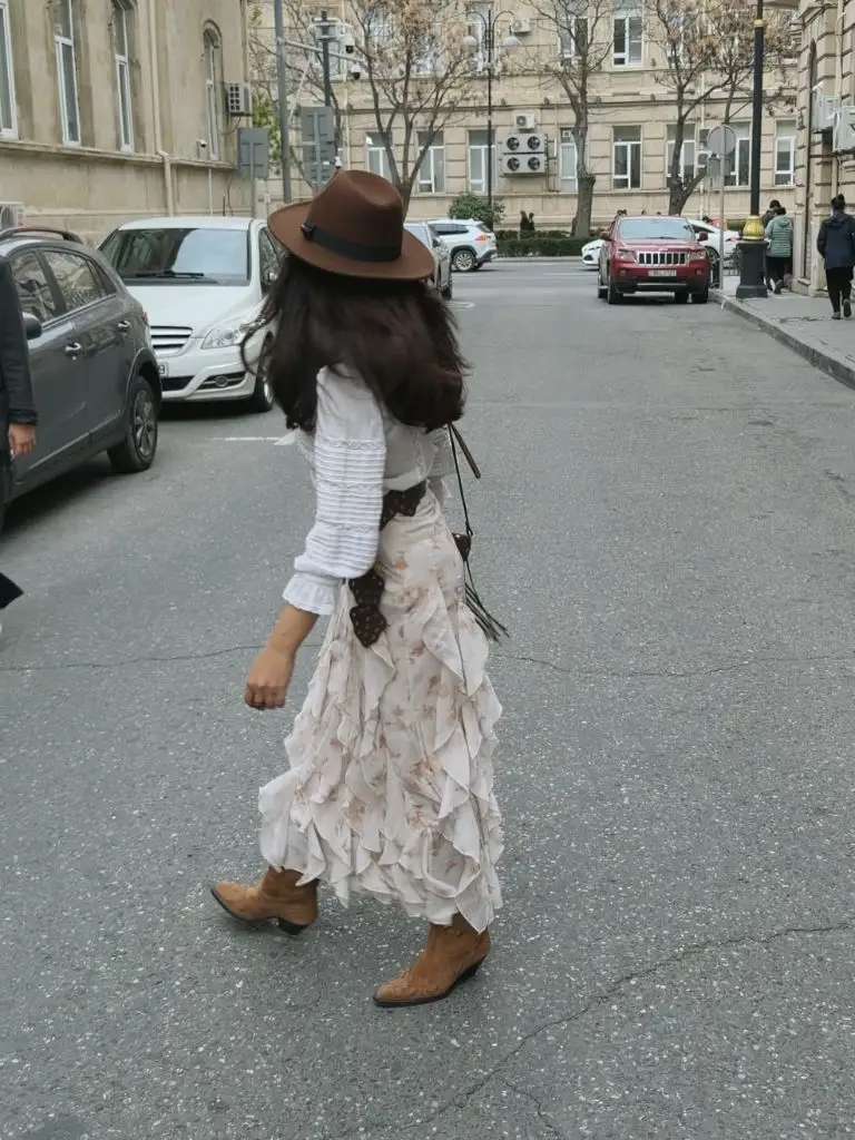 Walking shot of ruffled floral skirt with white blouse, cowboy boots, brown hat, and crossbody bag