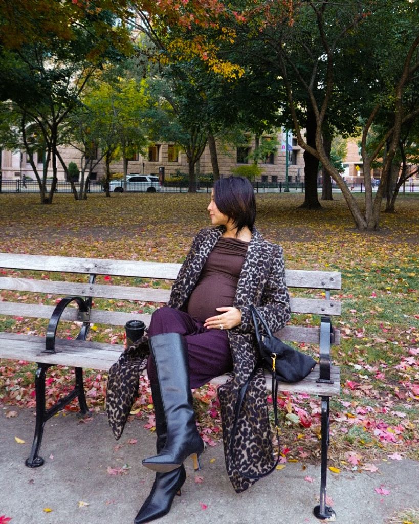 Pregnant woman on park bench wearing burgundy outfit, leopard coat, and tall black leather boots