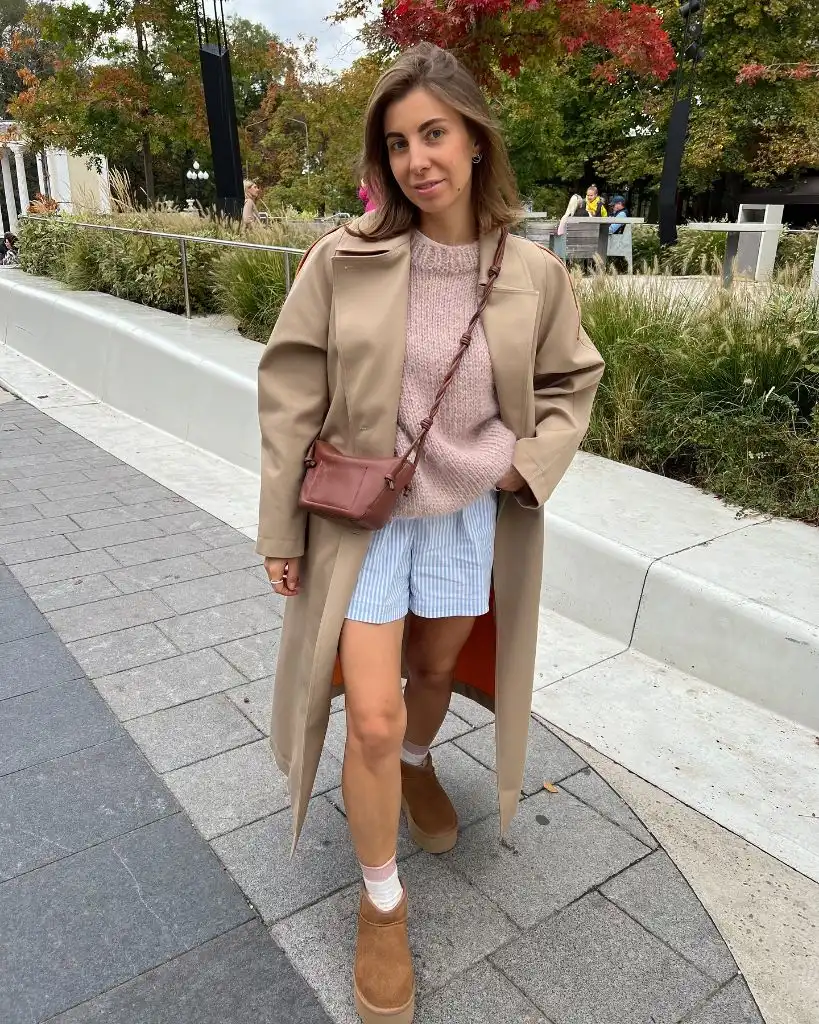 Beige trench coat over pink chunky knit sweater with blue striped shorts and UGG boots