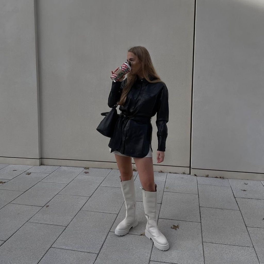 Black belted leather shirt dress over white mini skirt with white knee-high boots and black bag