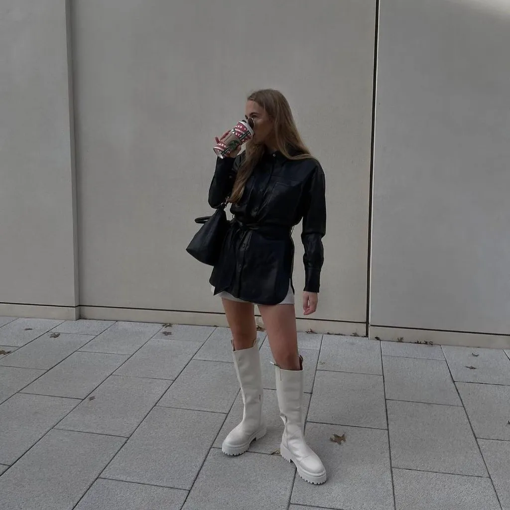 Black belted leather shirt dress over white mini skirt with white knee-high boots and black bag