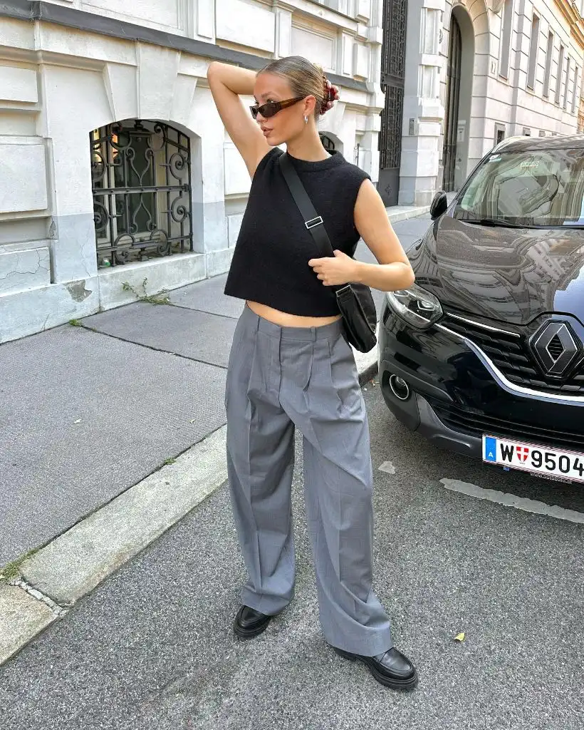 Black sleeveless crop top with grey pleated wide-leg trousers, black loafers, and black shoulder bag in Vienna