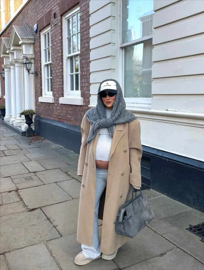 Pregnant woman in camel trench coat, gray hooded scarf, gray sweats, baseball cap, and UGG slides