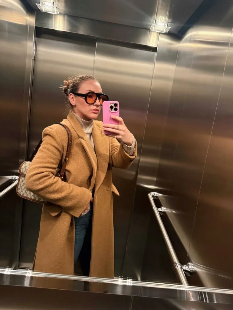 Camel wool coat with cream turtleneck, jeans, and Gucci bag in elevator mirror selfie