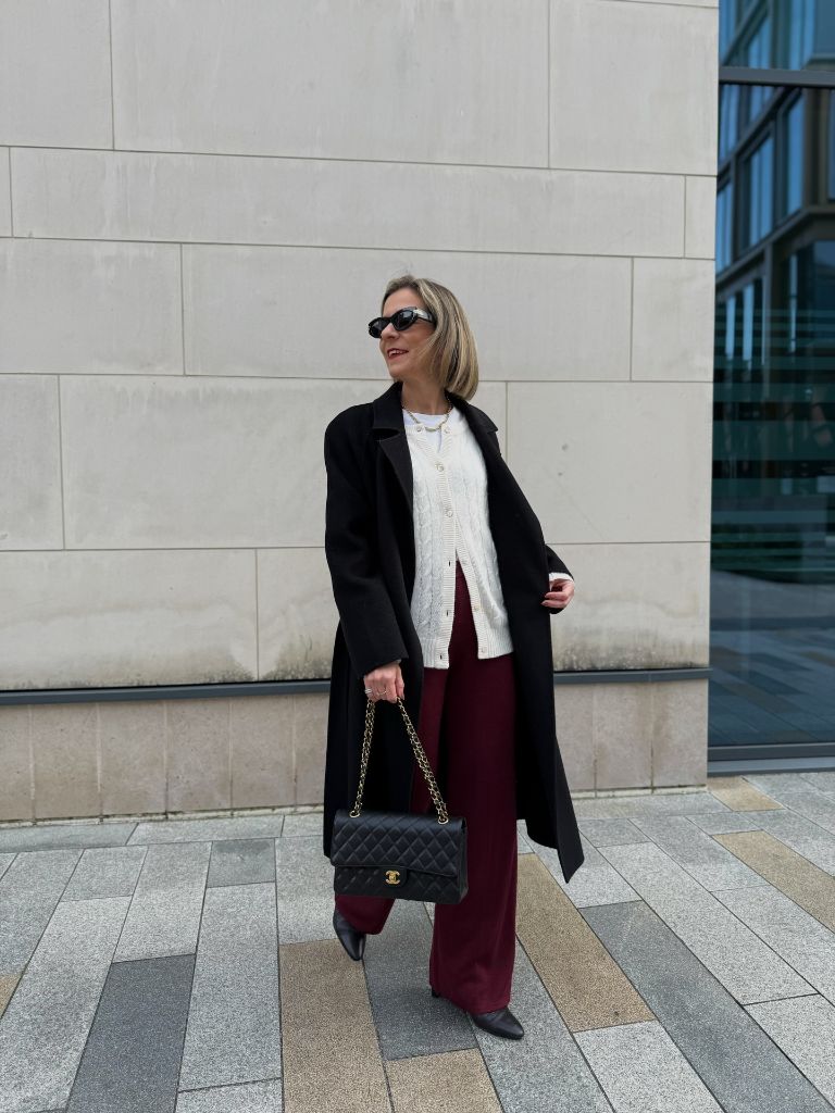 Black long coat over white cable knit cardigan and burgundy wide-leg trousers with Chanel bag