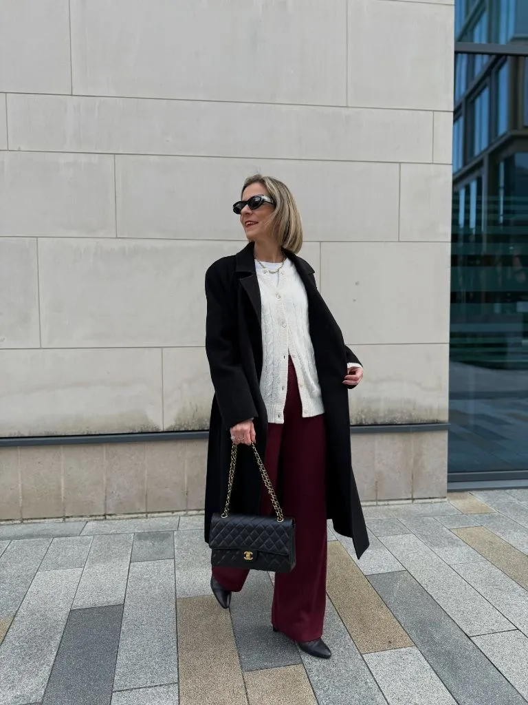 Black long coat over white cable knit cardigan and burgundy wide-leg trousers with Chanel bag