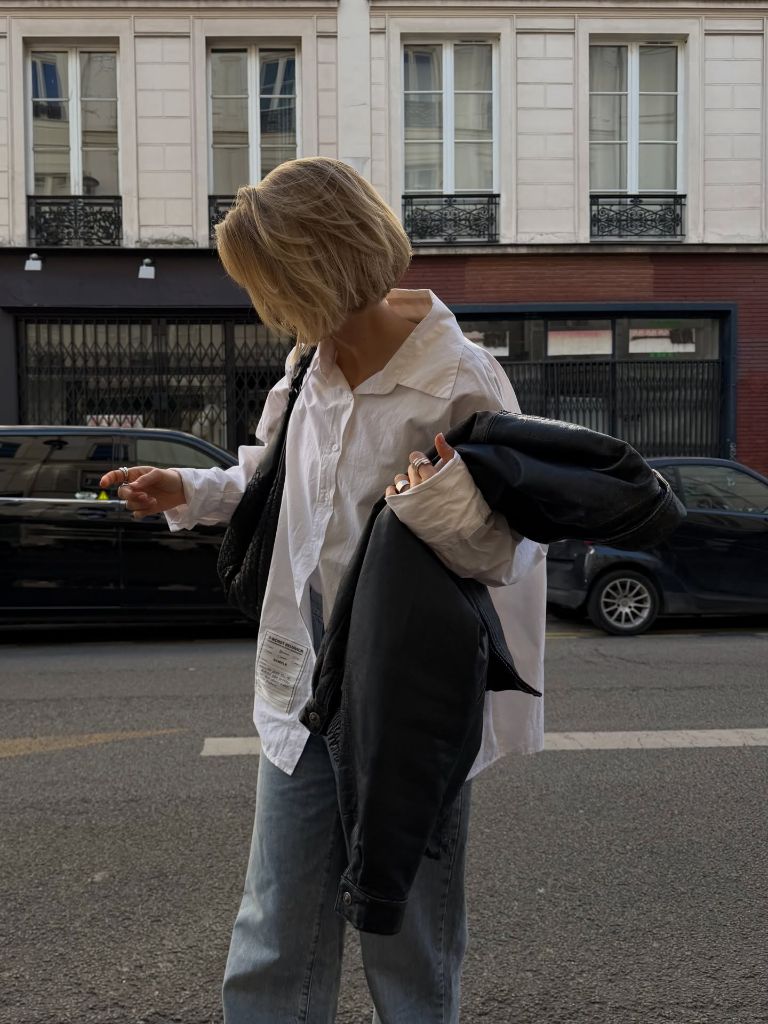 Oversized white button-up shirt with grey jeans and a black leather jacket over the arm on a Paris street