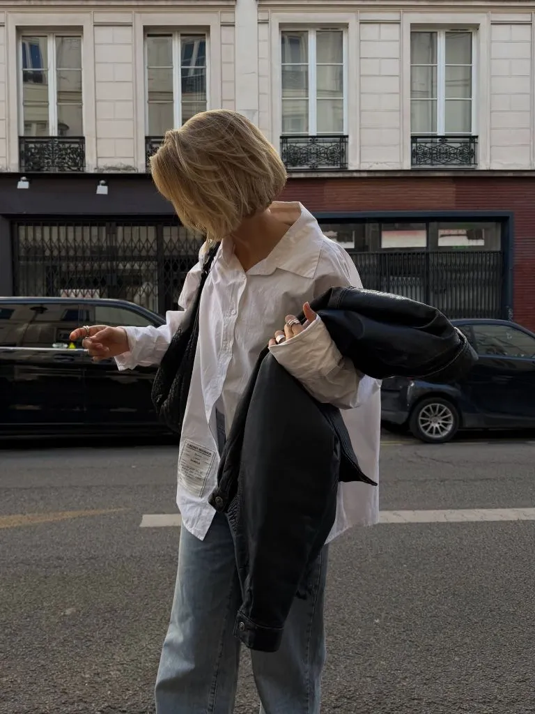 Oversized white button-up shirt with grey jeans and a black leather jacket over the arm on a Paris street