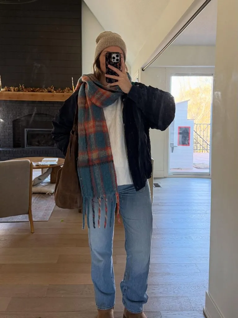 Mirror selfie of pregnant woman in dark jacket, white sweater, plaid scarf, beanie, and jeans with brown suede bag