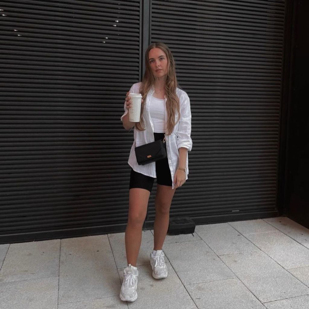 White oversized linen shirt over white tank top with black bike shorts, black crossbody bag, and white chunky sneakers