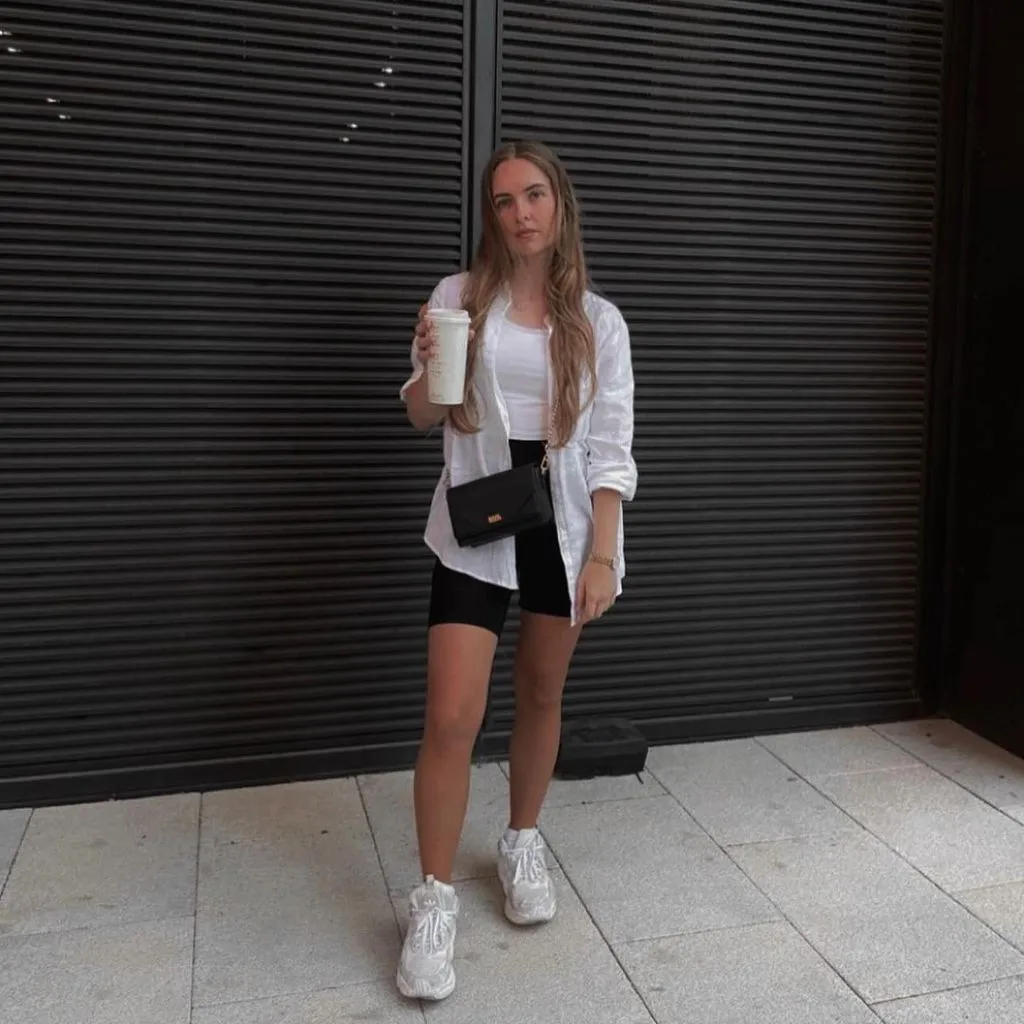 White oversized linen shirt over white tank top with black bike shorts, black crossbody bag, and white chunky sneakers