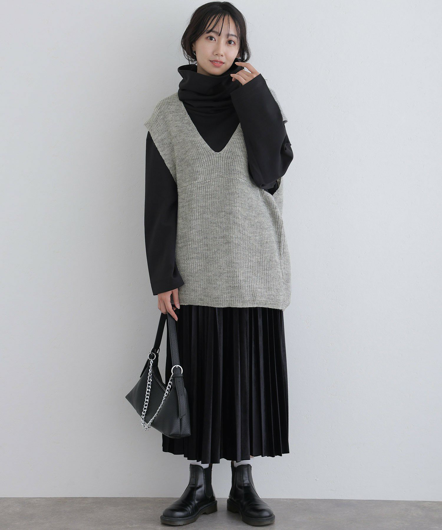 Woman in a grey knit vest over a black turtleneck with a black pleated midi skirt and black Chelsea boots