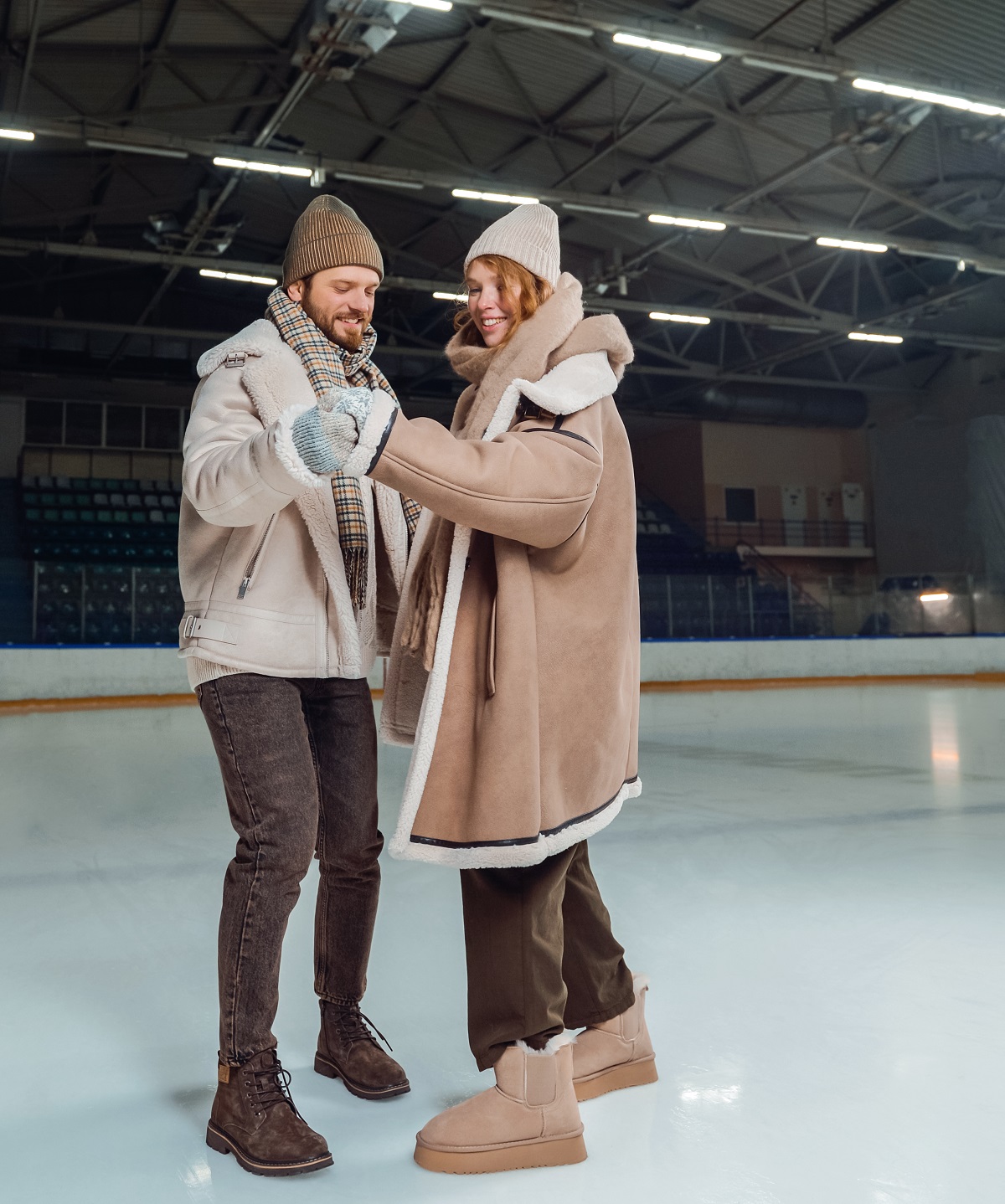 Couple on an indoor ice rink, the woman in a beige shearling coat with a knit hat, scarf and beige boots