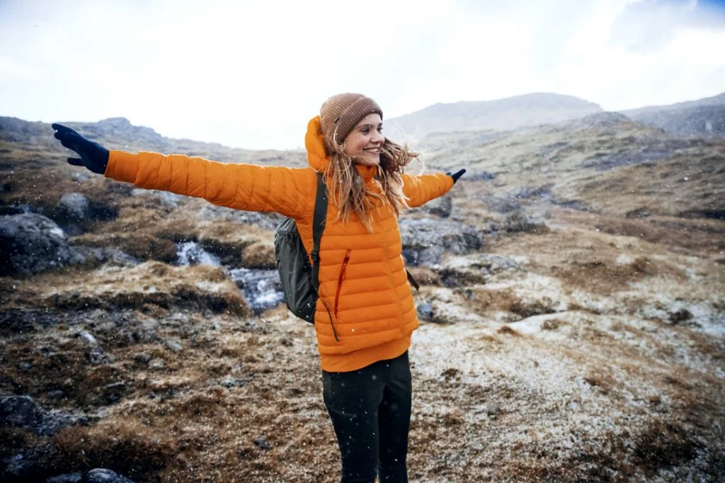 Woman in an orange puffer jacket with arms outstretched on a rocky mountain trail