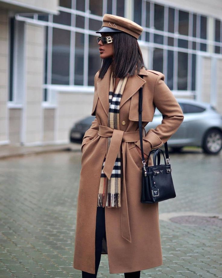 Woman outdoors wearing a camel belted wool coat, a check scarf, a baker boy cap and carrying a black handbag