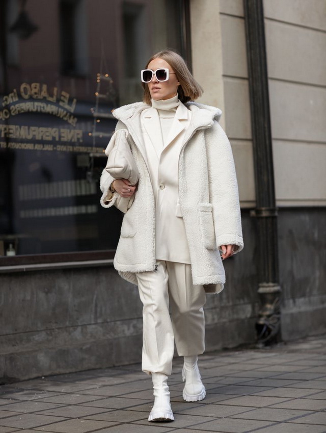 Woman wearing a cream teddy jacket layered over a white blazer and turtleneck with ivory trousers and white boots