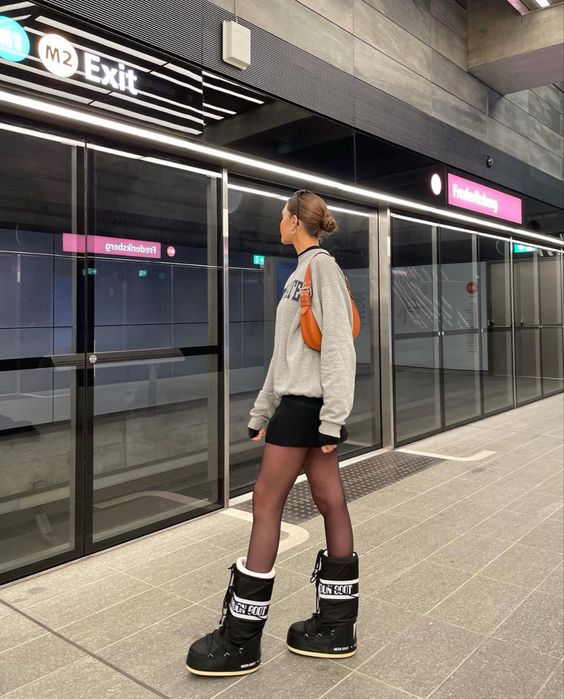 Woman in black Moon Boot Icon boots with logo band, grey sweatshirt, black mini skirt and sheer tights at a subway station