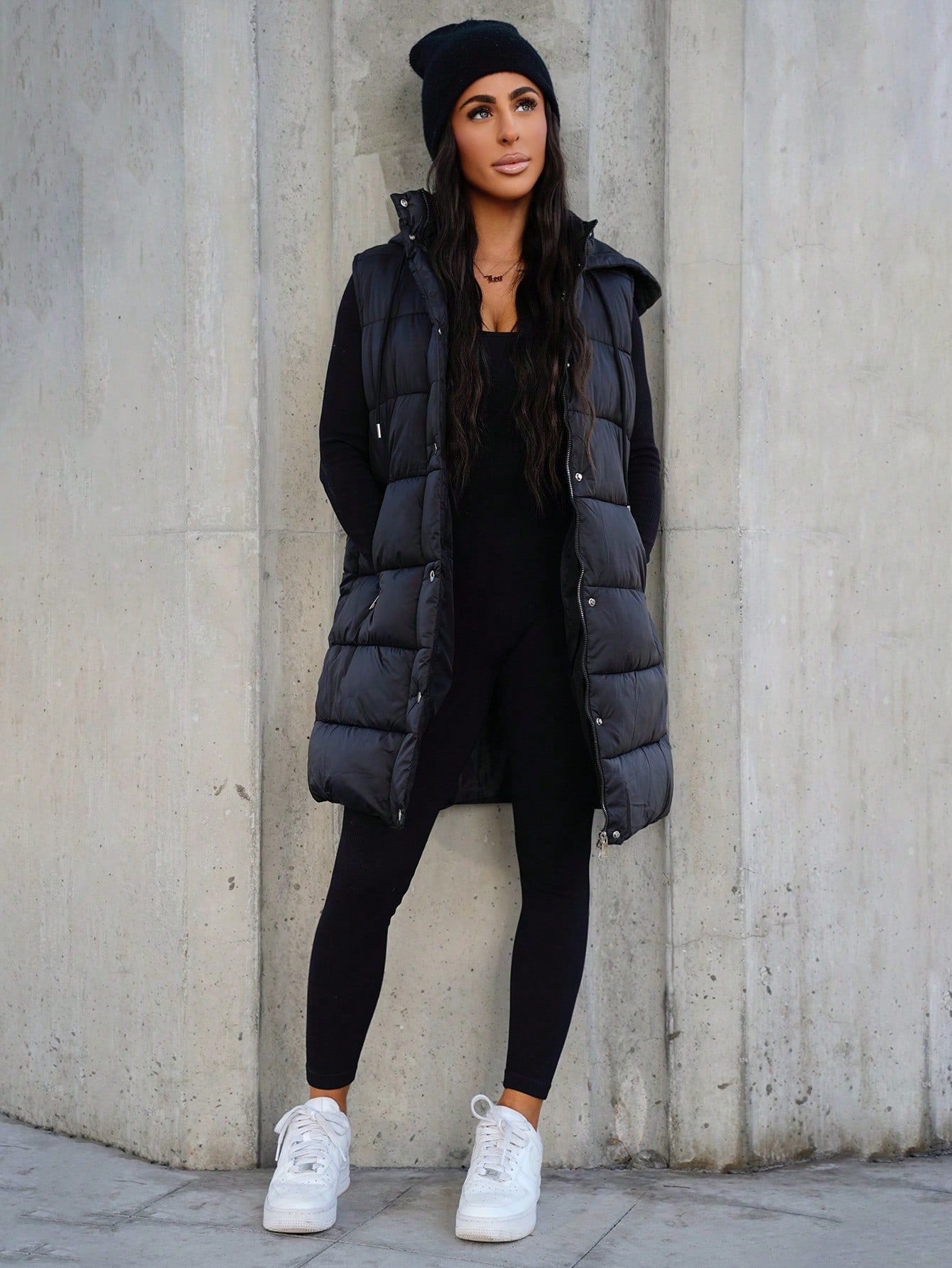 Woman wearing a long black puffer vest over black leggings with white sneakers and a black beanie