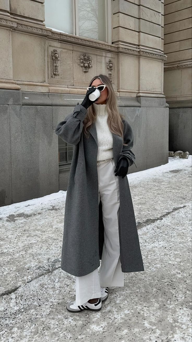 Woman wearing a grey oversized wool coat with cream knit sweater, white wide-leg trousers and sneakers in the snow