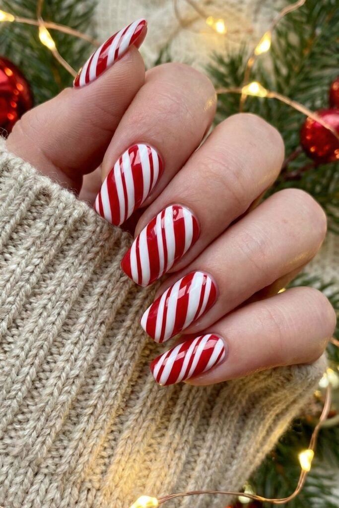 Almond-shaped nails with red-and-white candy-cane stripes, photographed against a Christmas tree with fairy lights; the hand wears a cream knit sleeve. Real photo, no watermark.