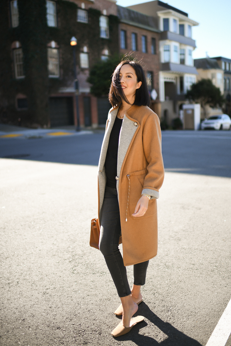 Woman in a camel coat with a black top, dark skinny jeans, camel mules and a tan bag