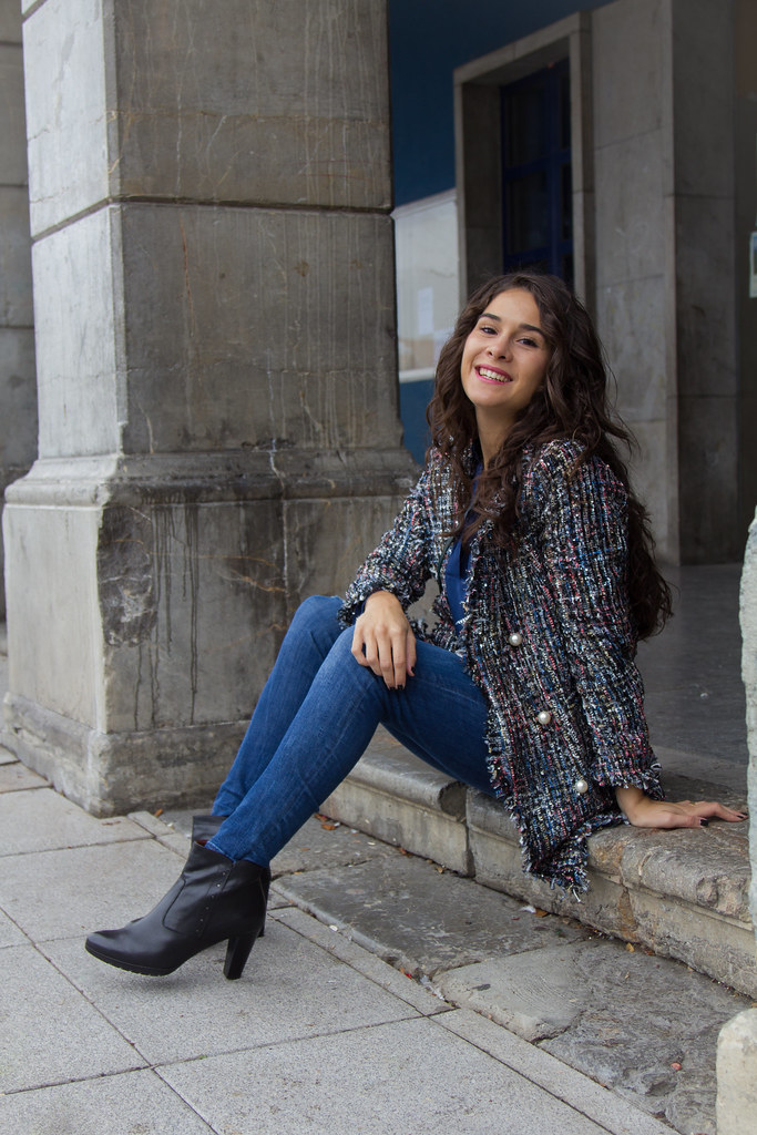Woman in a tweed jacket with pearl buttons, skinny jeans, and black ankle boots sitting on stone steps
