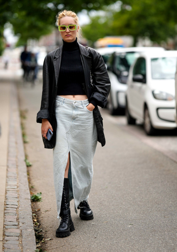 Woman in black platform lace-up combat boots, a light-wash denim maxi skirt with a front slit, a black turtleneck and a black leather jacket