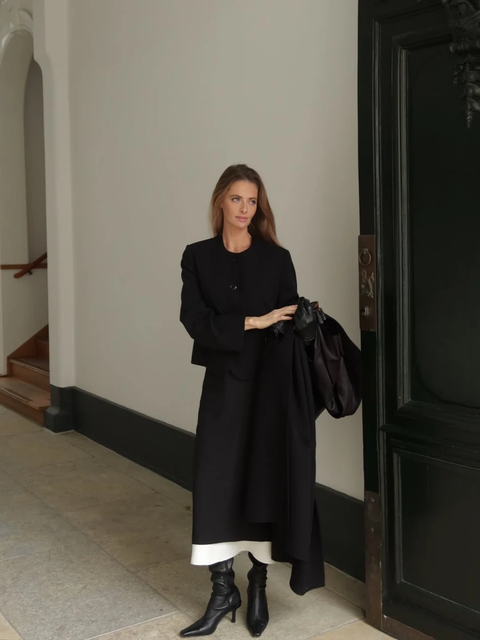 Woman in an all-black outfit with a coat draped over her arm and heeled boots