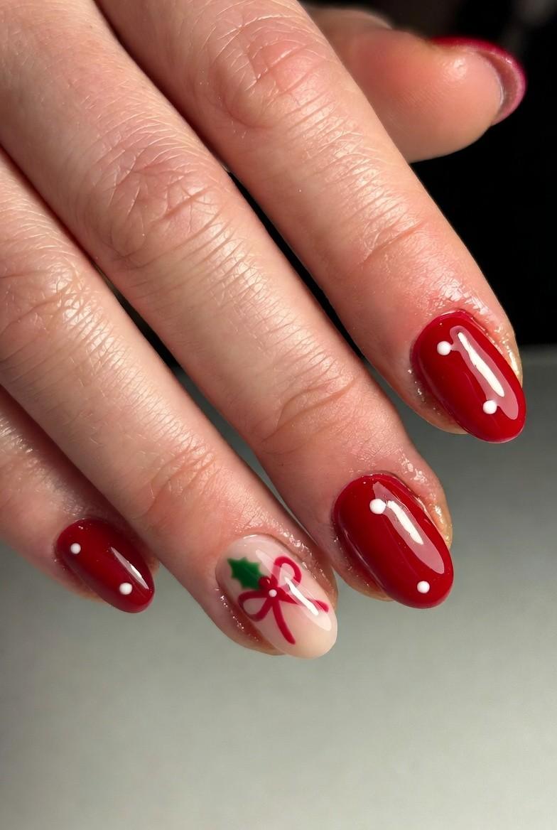 Glossy cherry-red oval nails with white dot accents and a nude accent nail with a red bow and holly leaf