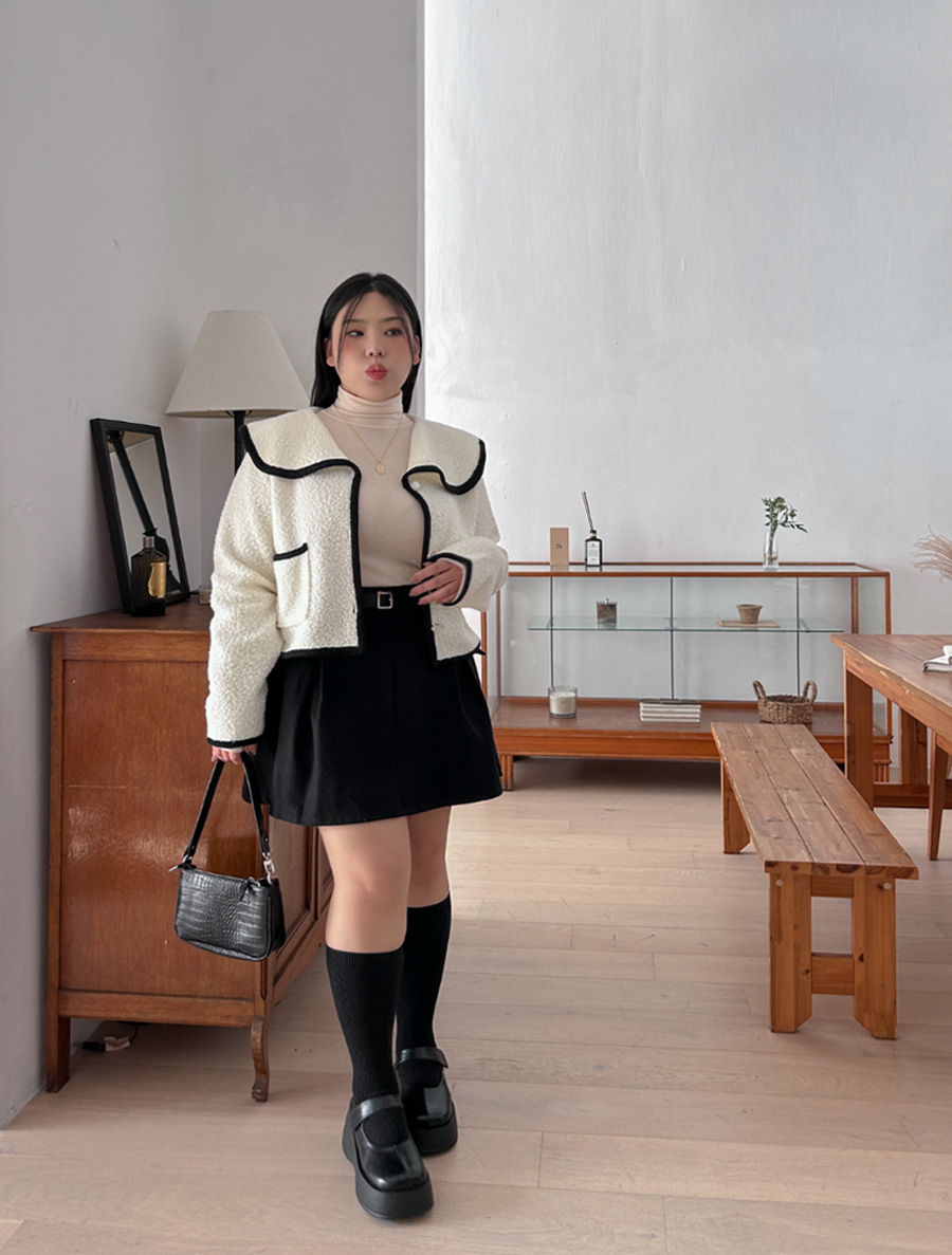 Woman in a cropped cream teddy jacket with black trim, black mini skirt, knee socks and platform Mary Janes