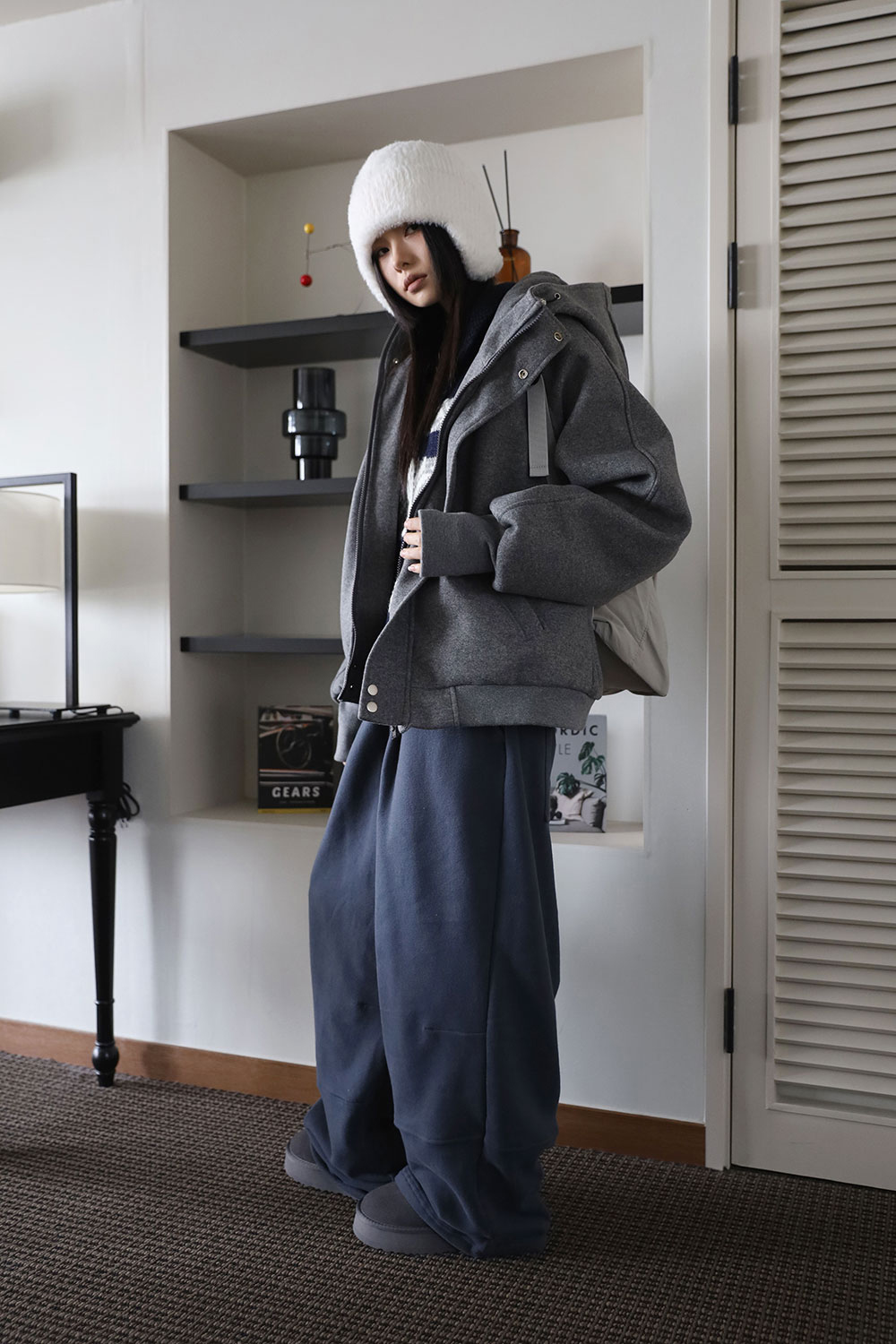 Woman in an oversized grey hooded jacket, white fuzzy trapper hat, navy wide sweatpants and grey slipper boots