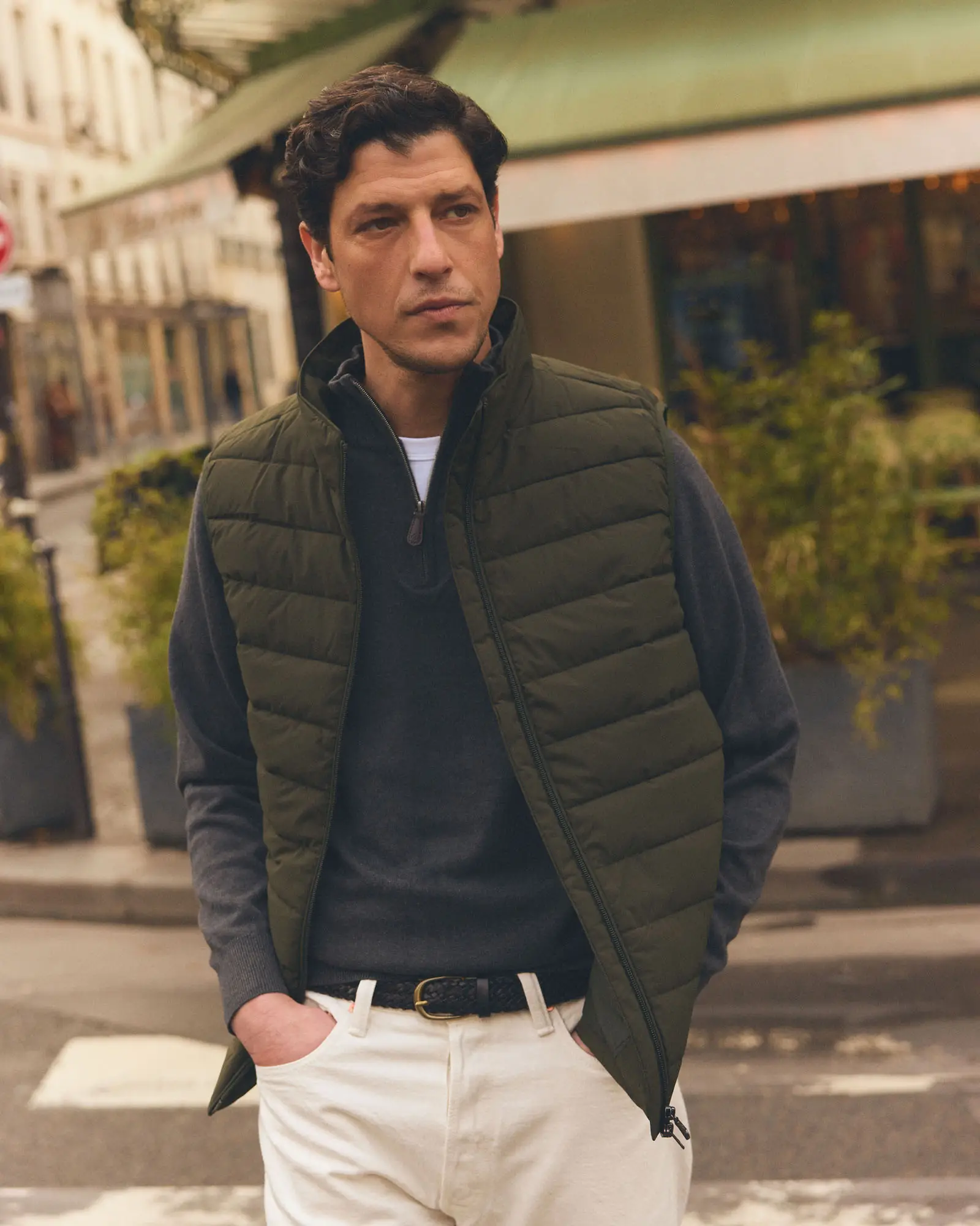 Man wearing an olive quilted gilet over a grey quarter-zip with white chinos on a city street