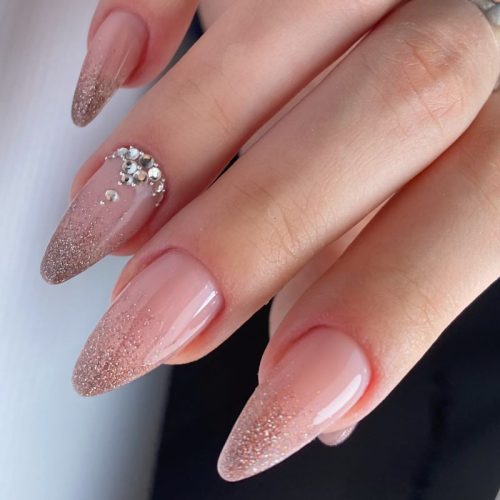 Nude almond nails with rose-gold glitter ombre fading from the tips and a crystal accent