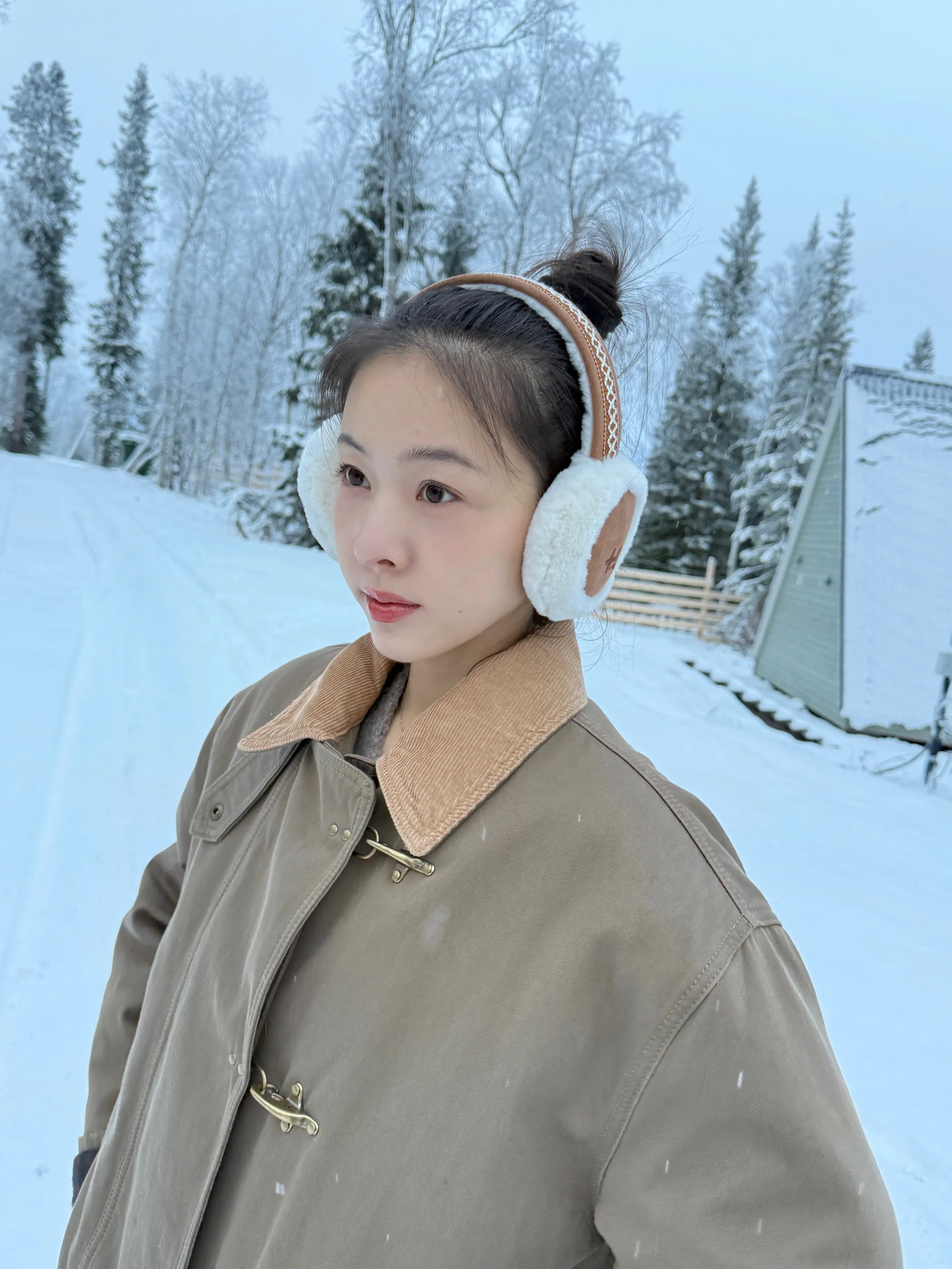Woman wearing white shearling earmuffs and an olive field jacket in a snowy forest