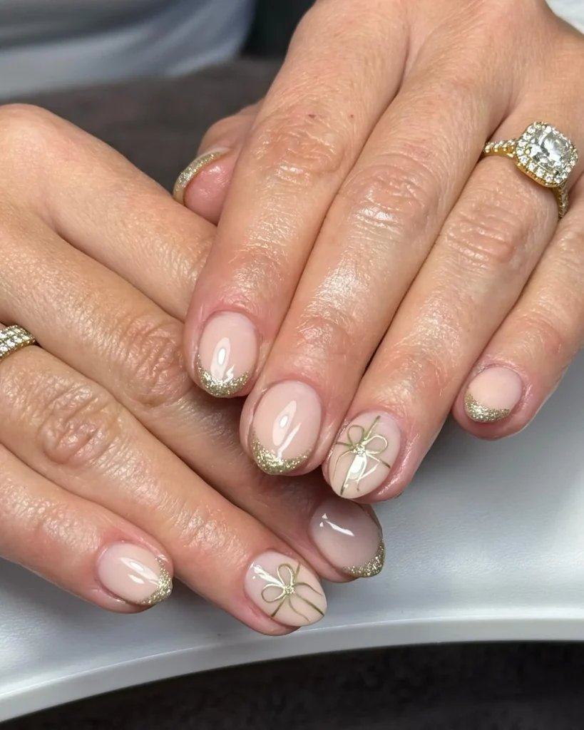 Short nude nails with gold glitter French tips and hand-drawn gold bow accents