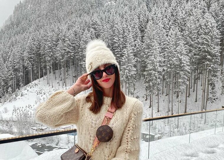 Woman in a white fur hat, cream chunky knit sweater and sunglasses in snowy mountains