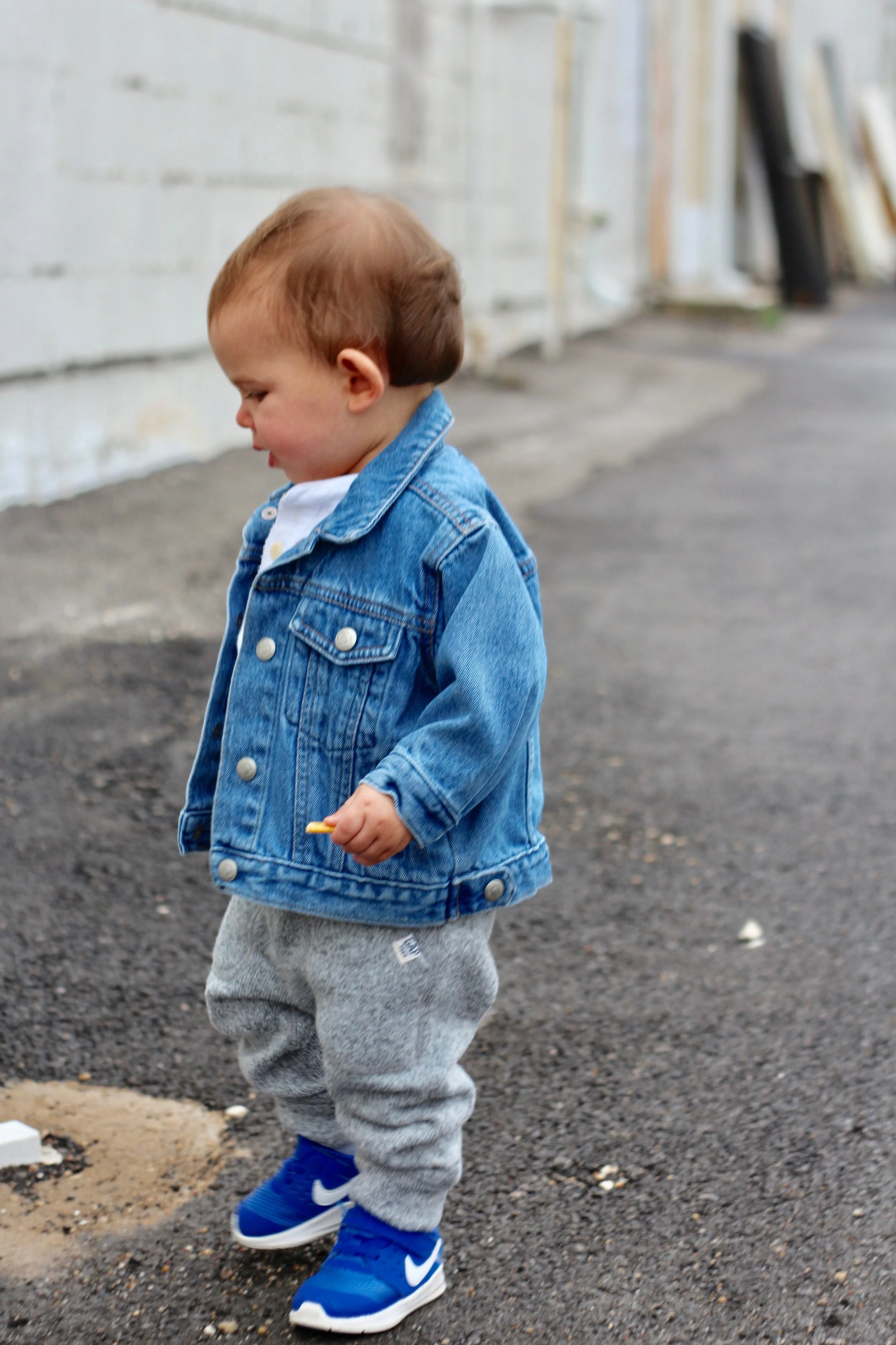 A toddler wearing a blue denim trucker jacket with gray sweatpants and blue sneakers