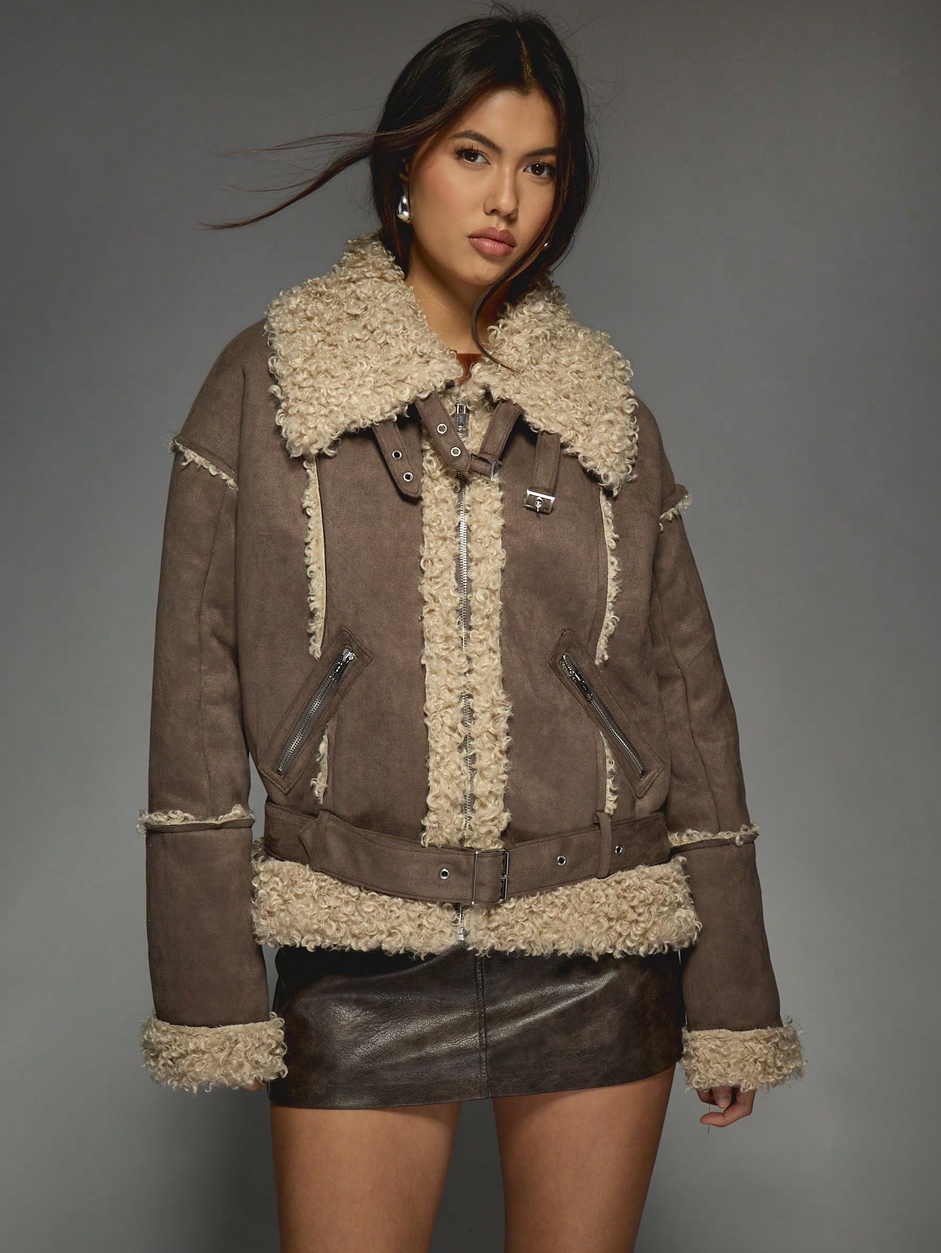 A brown shearling-lined aviator jacket with a curly cream collar, belted waist and zip pockets, worn with a dark leather mini skirt.