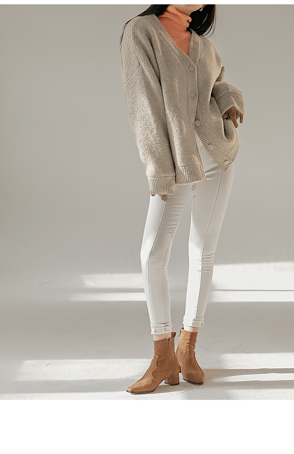 Woman wearing an oversized taupe V-neck cardigan over a peach turtleneck with white skinny jeans and tan ankle boots