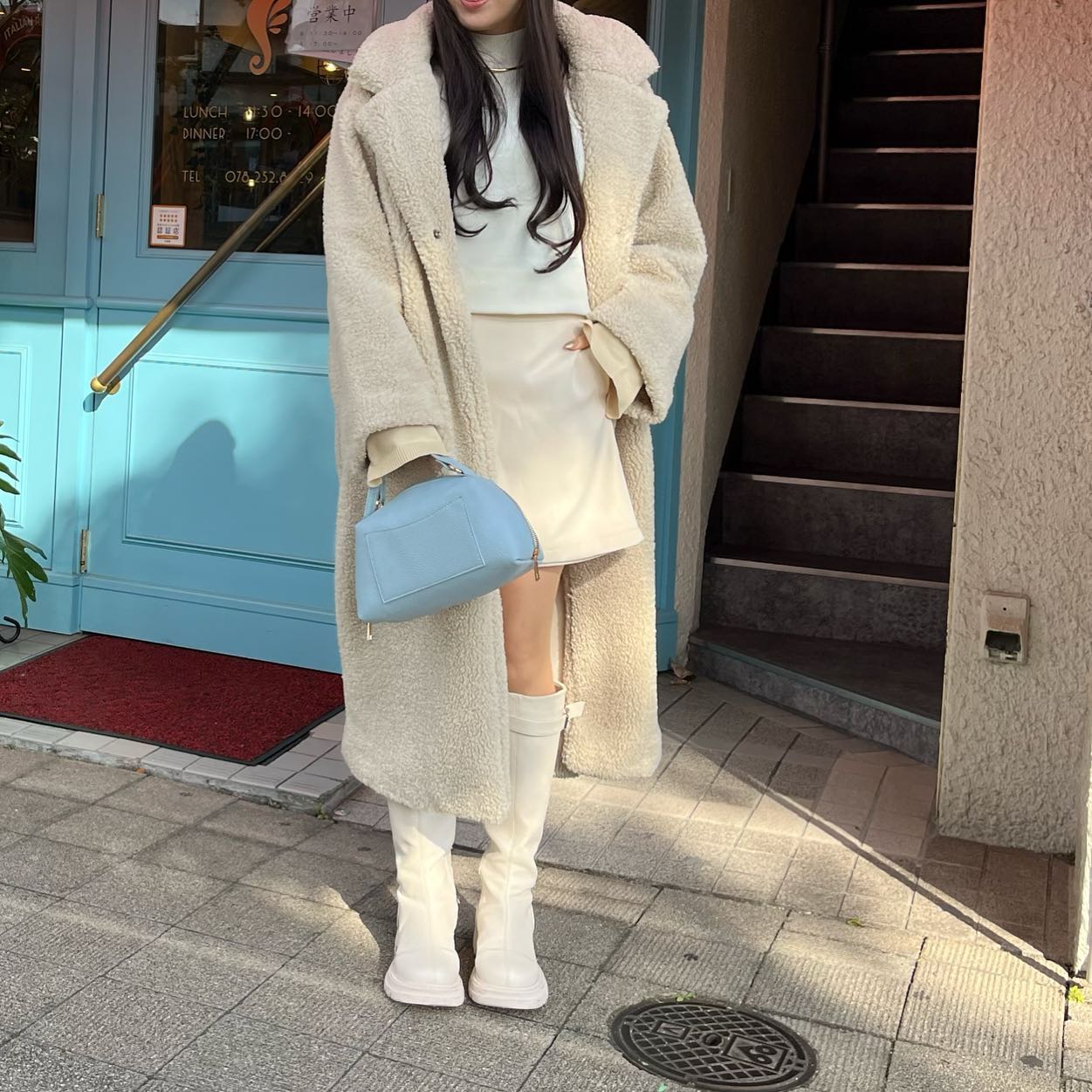 Woman in a long cream teddy coat with a cream mini skirt, white knee-high boots and a light blue handbag