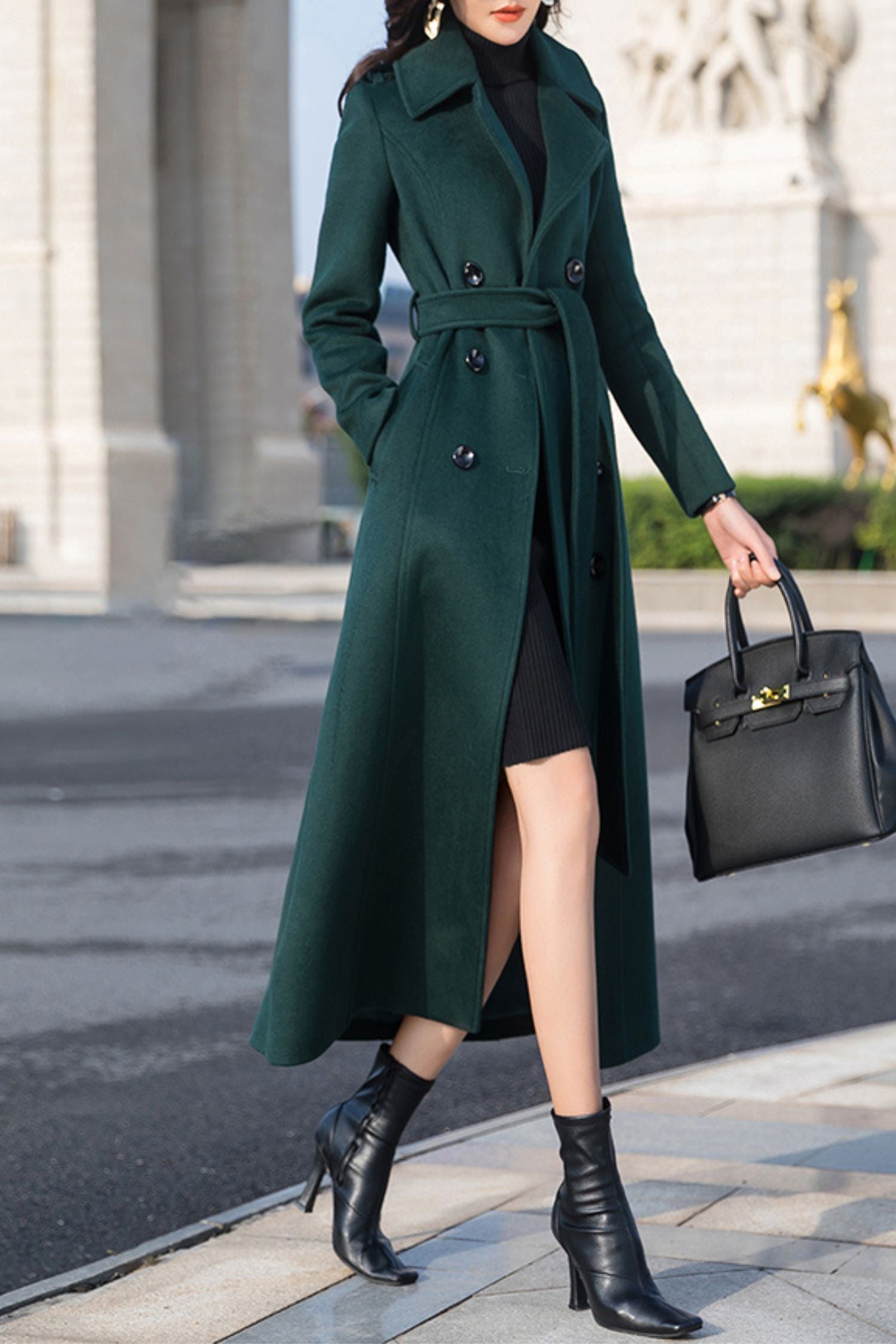 A dark emerald green double-breasted belted long coat over a black dress with black heeled boots and a black handbag.