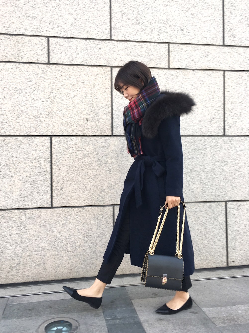 Woman wearing a navy belted coat with a tartan plaid scarf and black flats