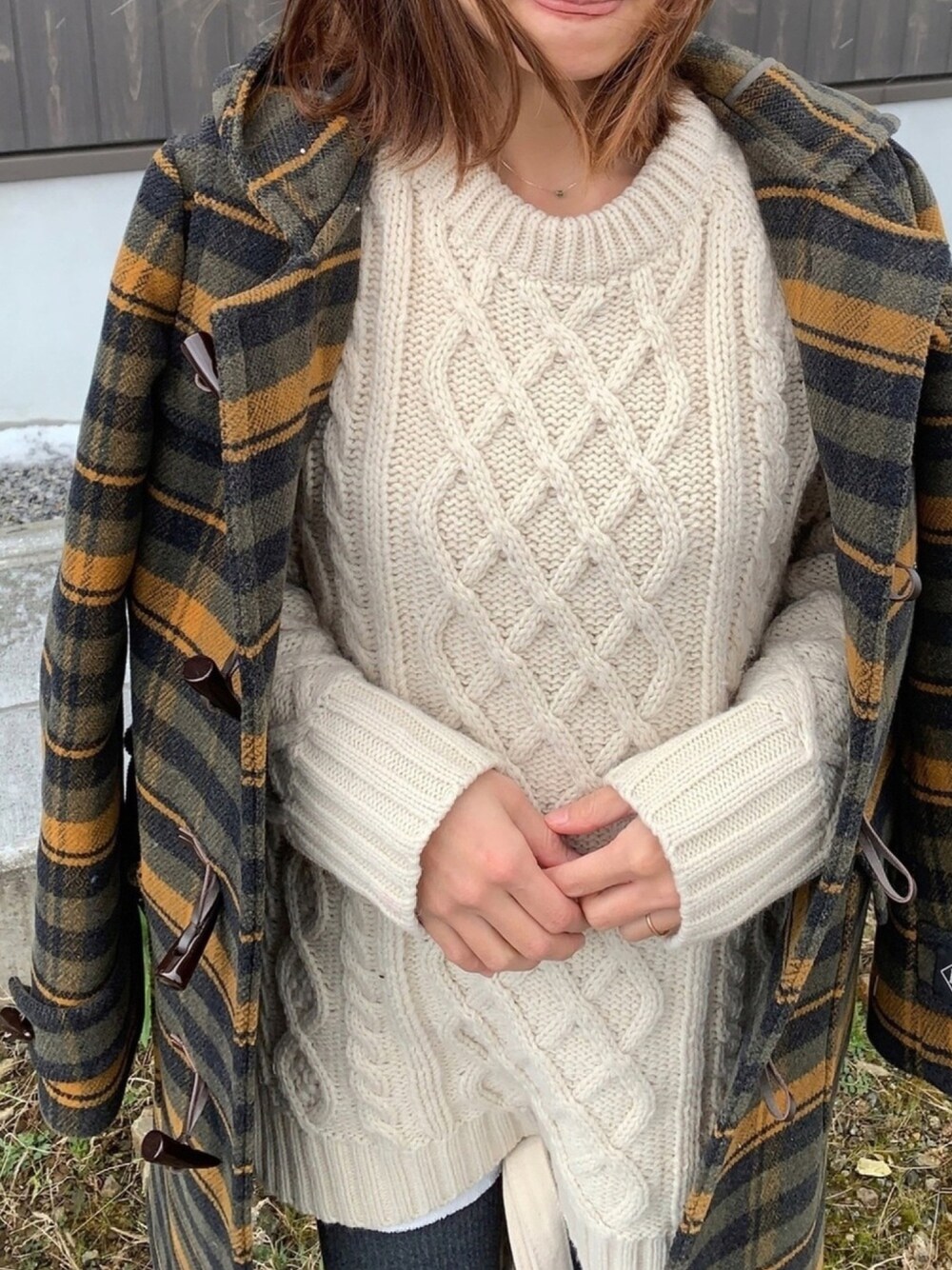 Close-up of a cream cable-knit sweater worn under an open brown-and-gold plaid toggle duffle coat
